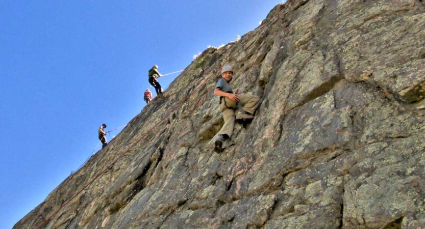People wearing safety gear are attached to ropes as they climb a rock wall under a blue sky